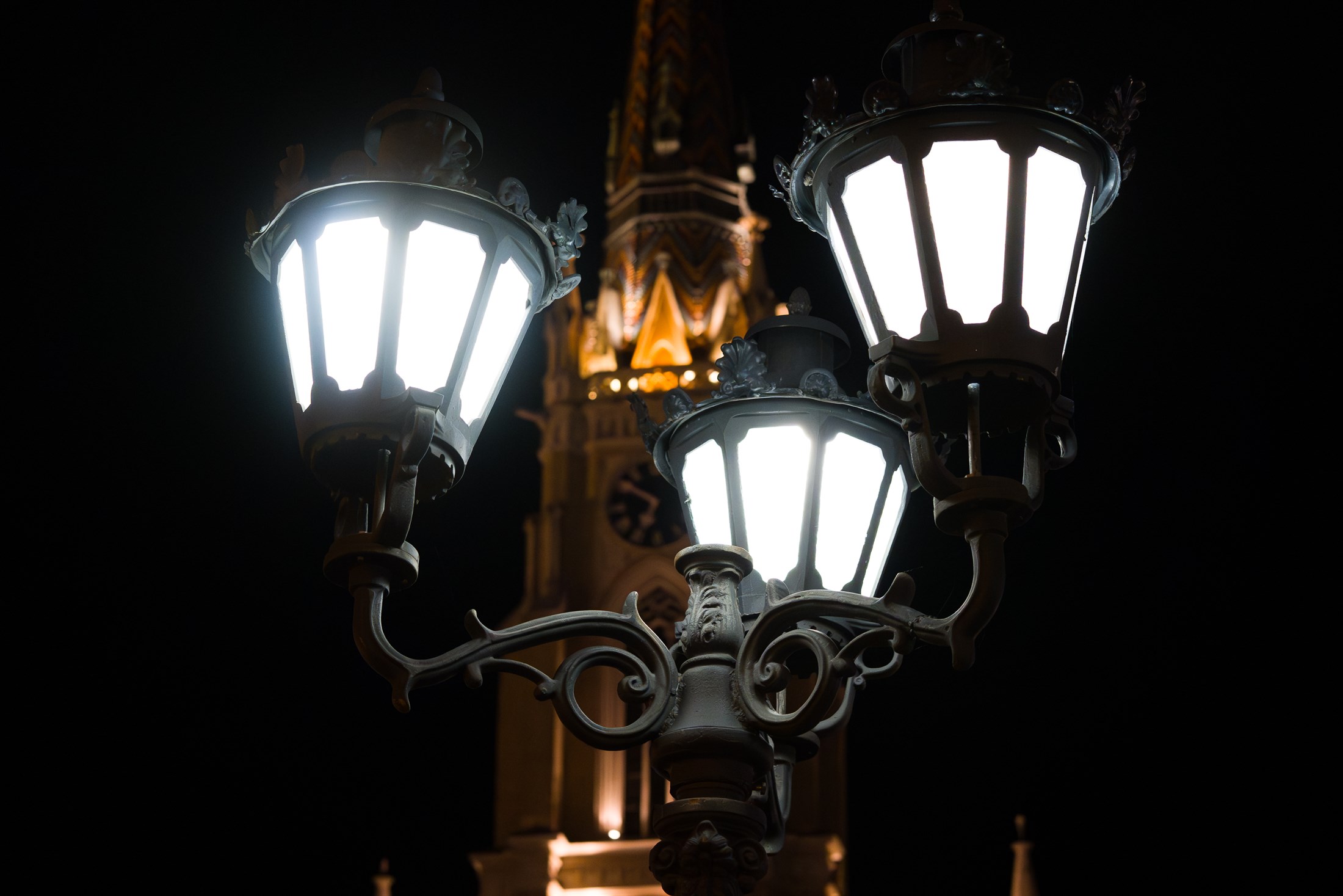 Three white street lamps burning at night, a floodlit church tower rising gold and out of focus behind them.