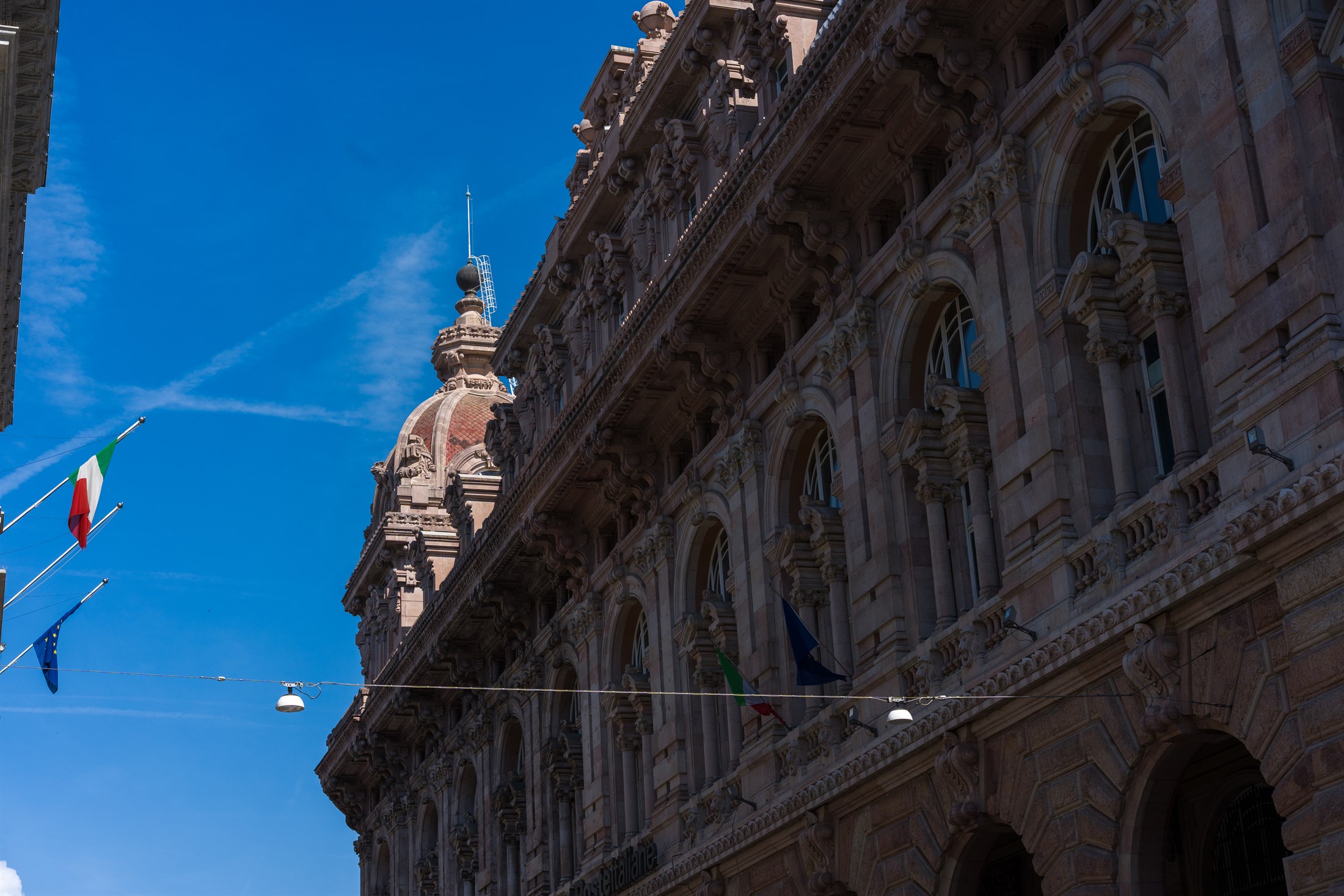 A grand stone palace front raking away against a deep blue sky, Italian and European flags hanging from its corner.