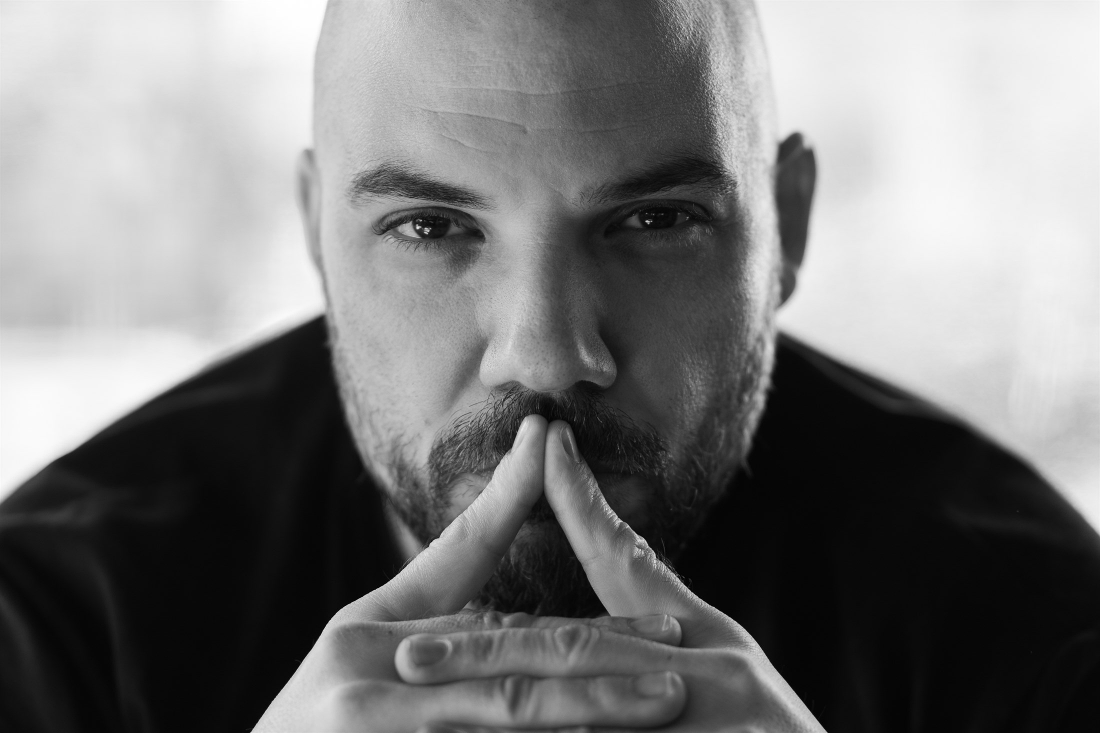 A close black and white portrait of a bearded man against a blown-out window, hands steepled against his mouth.