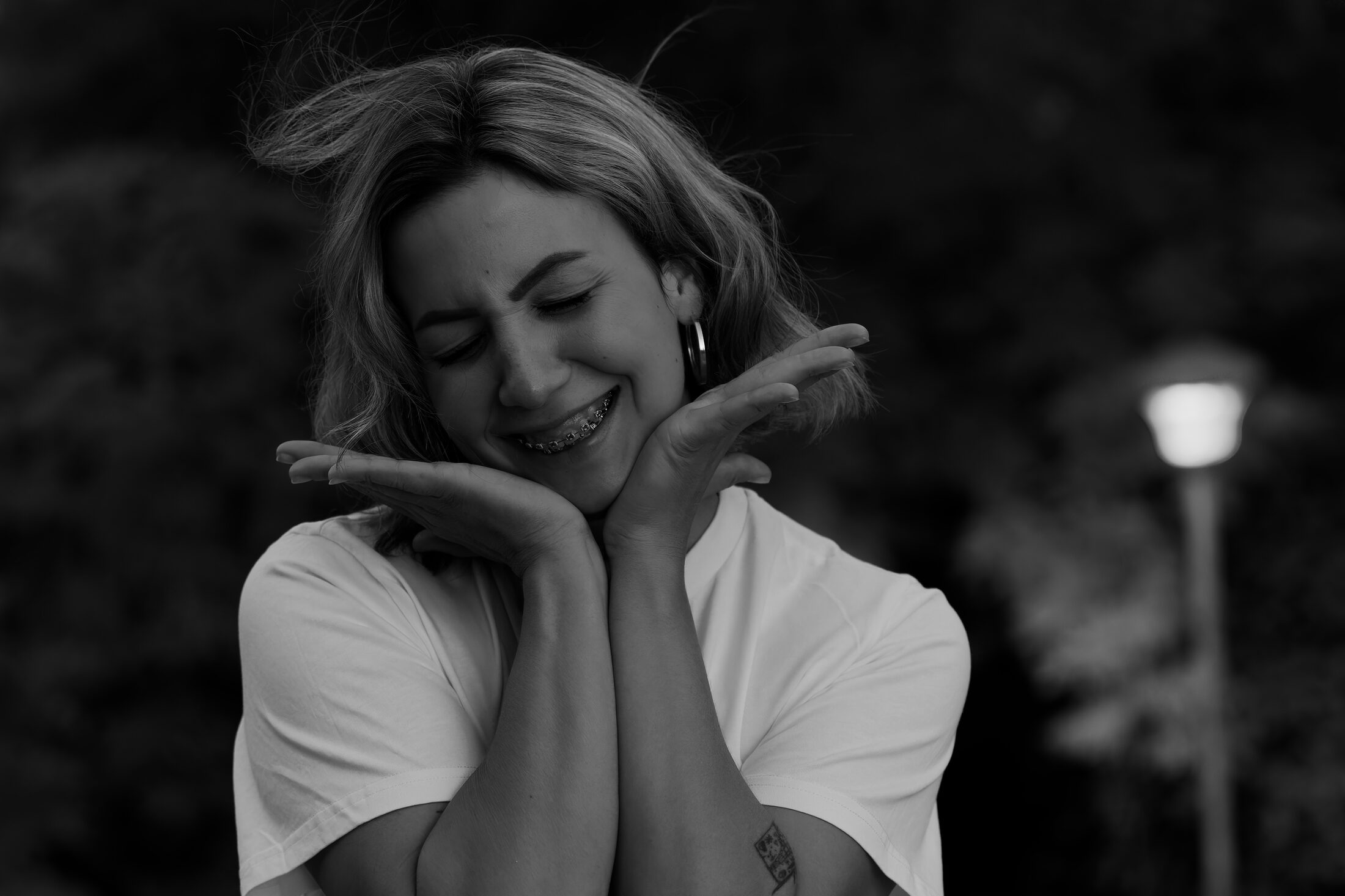 A young woman laughing with her eyes closed in black and white, both hands cupped under her chin, a garden lamp burning out of focus behind her.