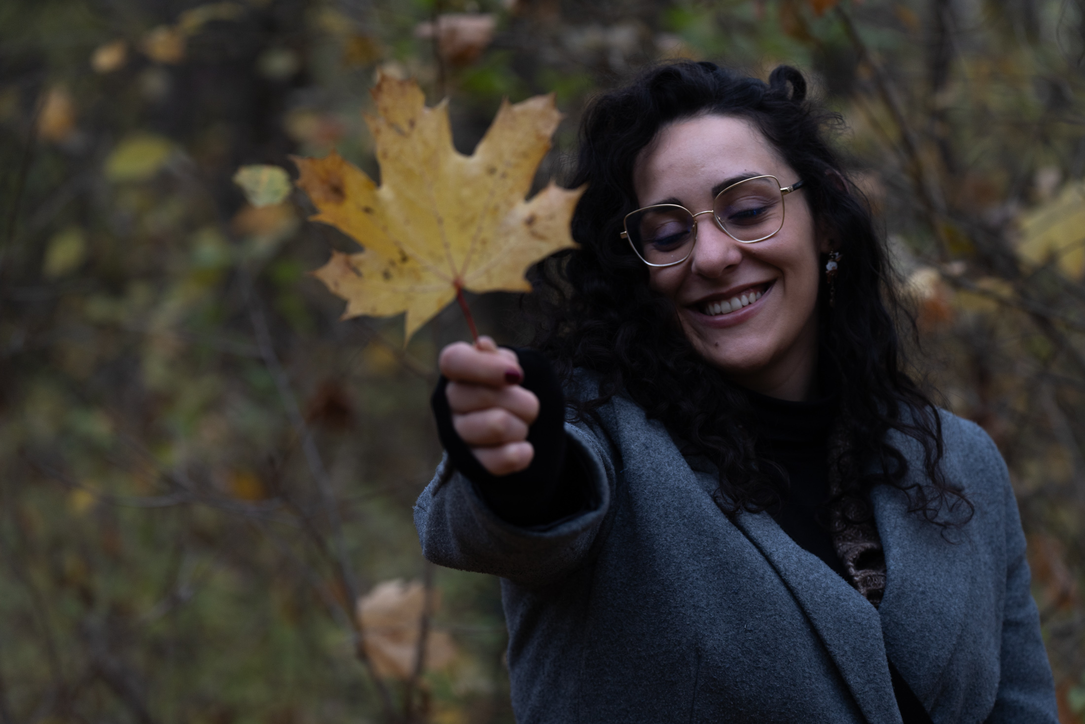A woman in a grey wool coat and glasses holding a yellow maple leaf out towards the camera, laughing, autumn woodland behind her.