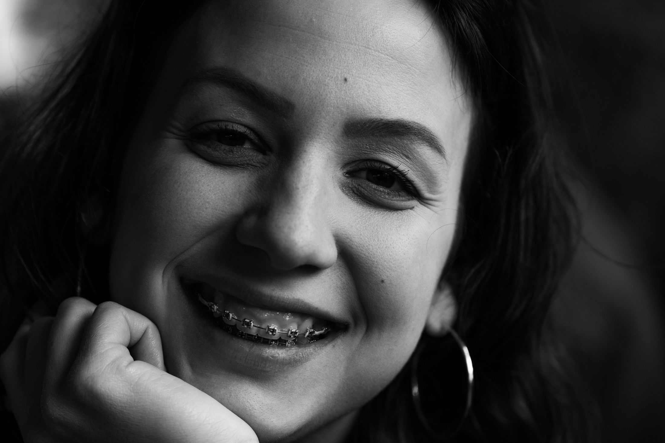 A close black and white portrait of a young woman laughing, chin resting on her hand, hoop earring catching the light.