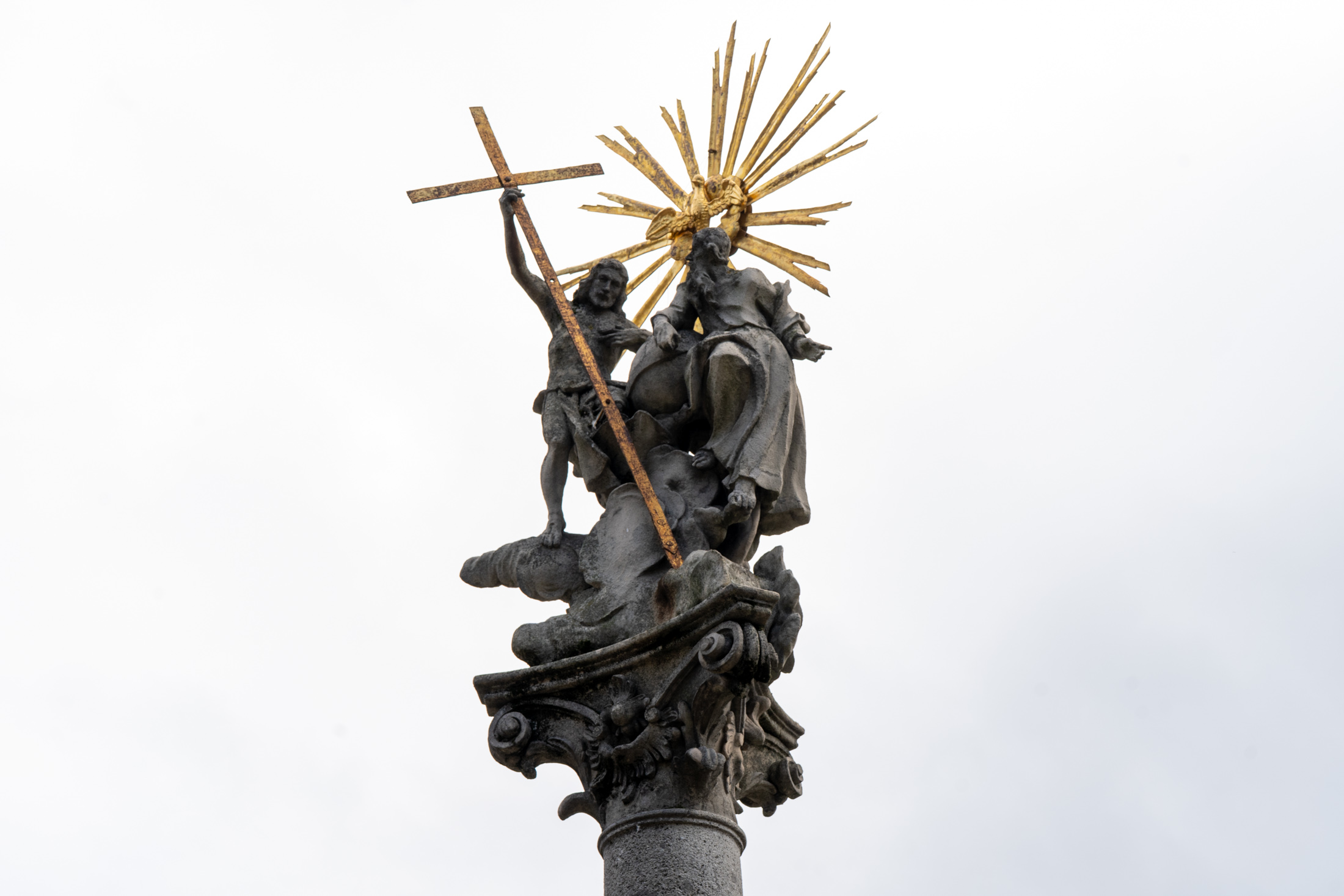 A baroque column crowned with a gilt cross and sunburst against a pale sky.
